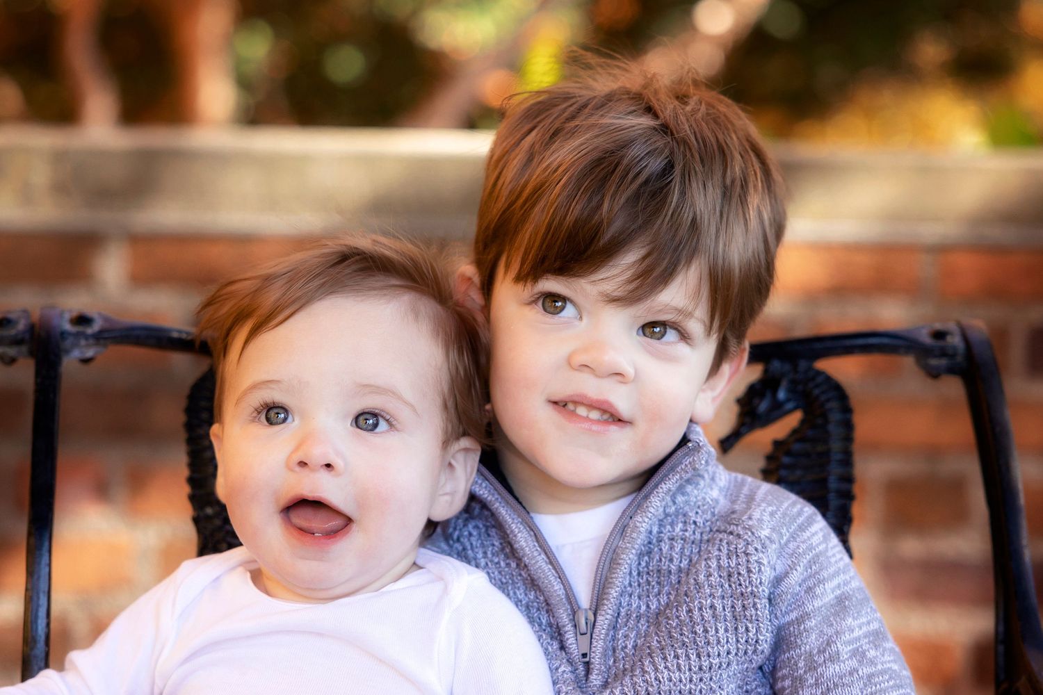Two young children sitting close together on outdoor furniture smiling at camera.