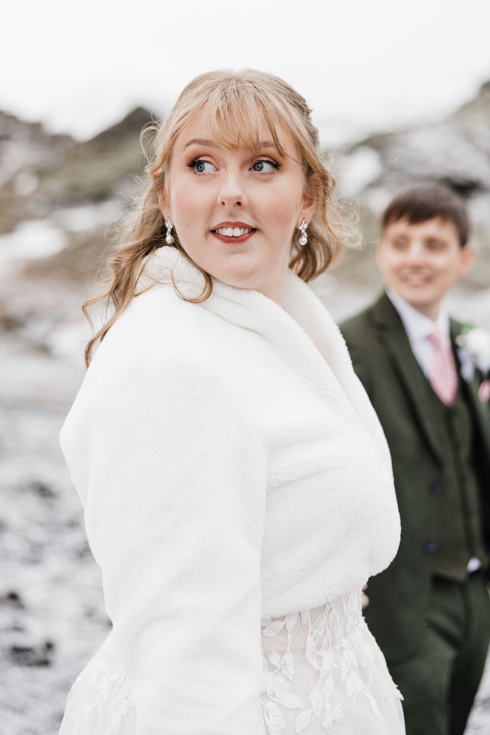 A wedding couple poses on a rocky beach with snowy mountains in the background.