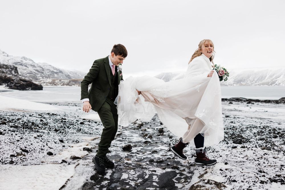 A playful wedding couple dances on rocky terrain with snow-covered mountains behind them.