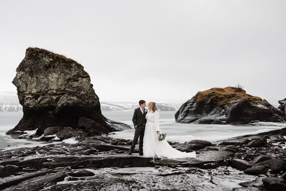 Wedding couple stands on dramatic coastal rocks with mountains and ocean in the background.