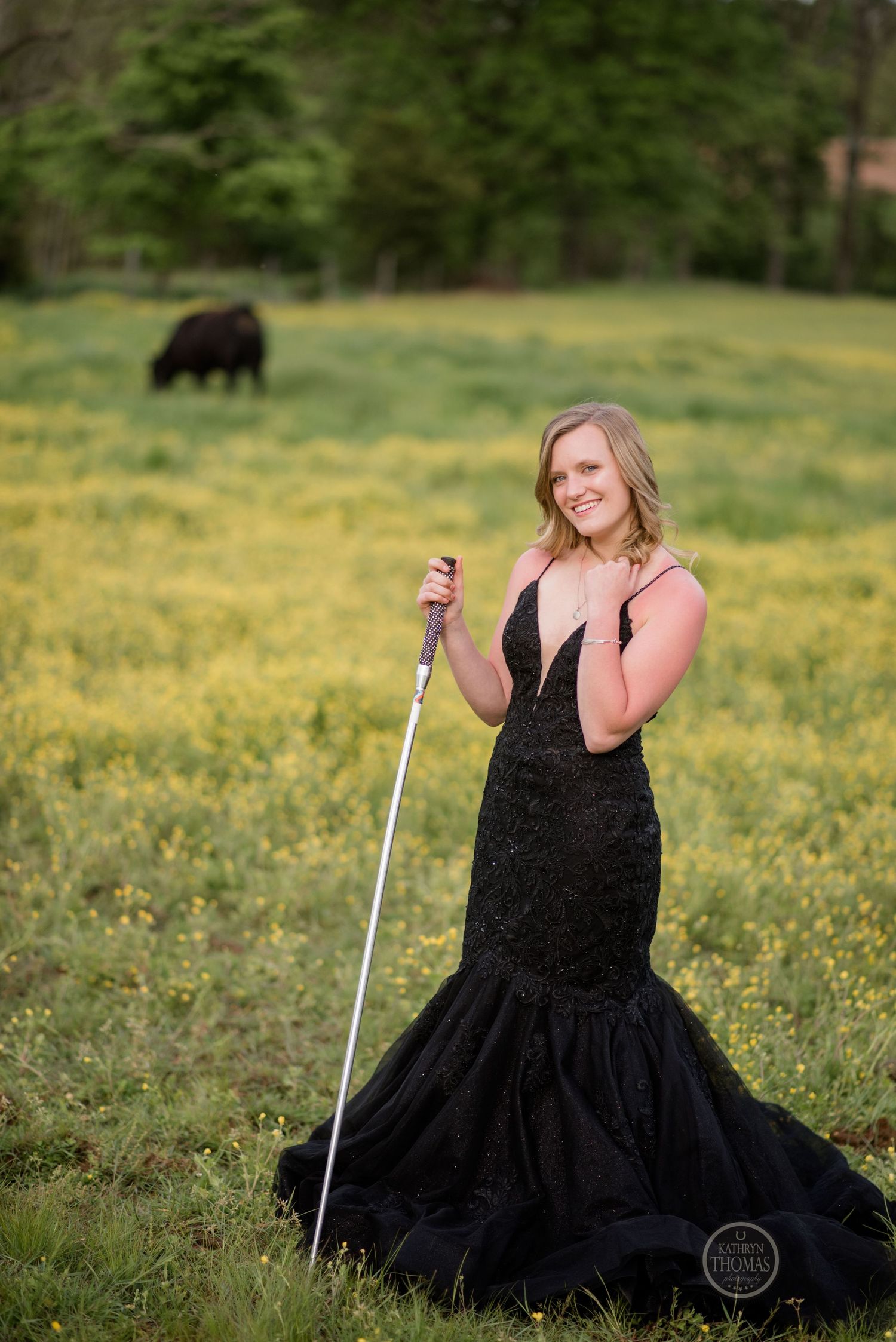 An elegant black dress stands out against a field of yellow flowers with a grazing cow in the distance.