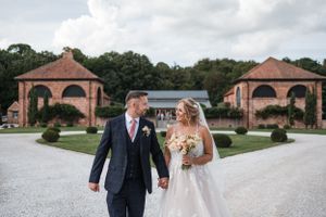 A couple walks along a gravel path between two brick gazebos at a formal garden venue on their wedding day at Hazel Gap Barn, Nottingham.