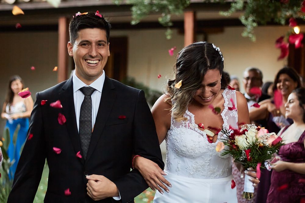 A newlywed couple walks down an aisle while guests shower them with red rose petals.