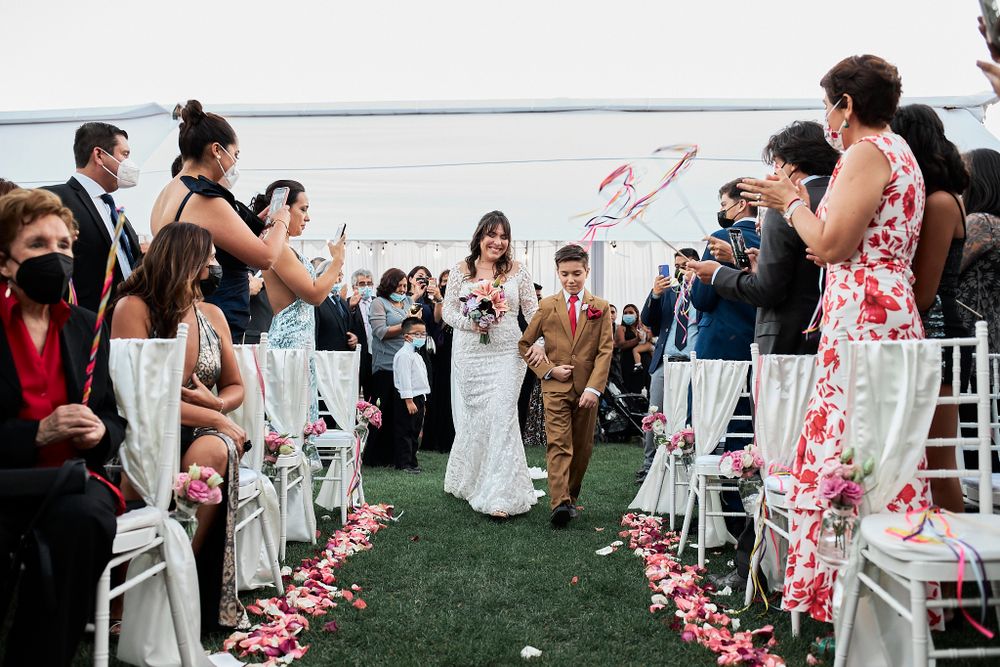 A couple walks down an outdoor aisle with white chairs and rose petals by the waterfront.