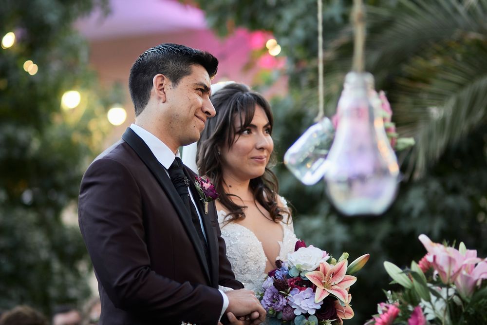 A couple shares an intimate moment in a garden setting with hanging lights and pink lilies.