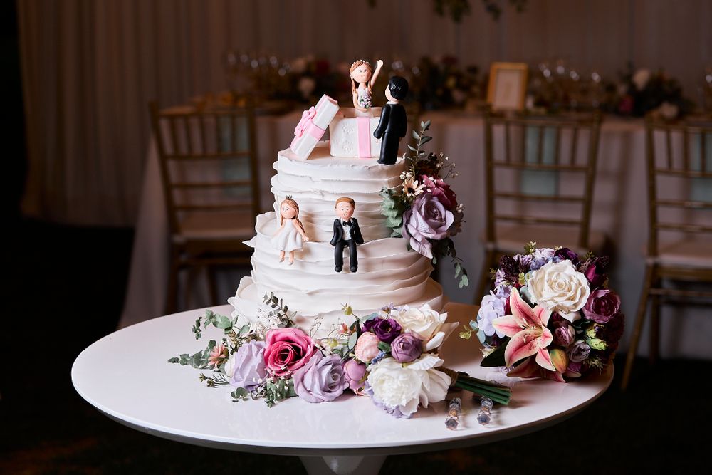 Wedding cake decorated with pink flowers and miniature couple topper on white display stand.
