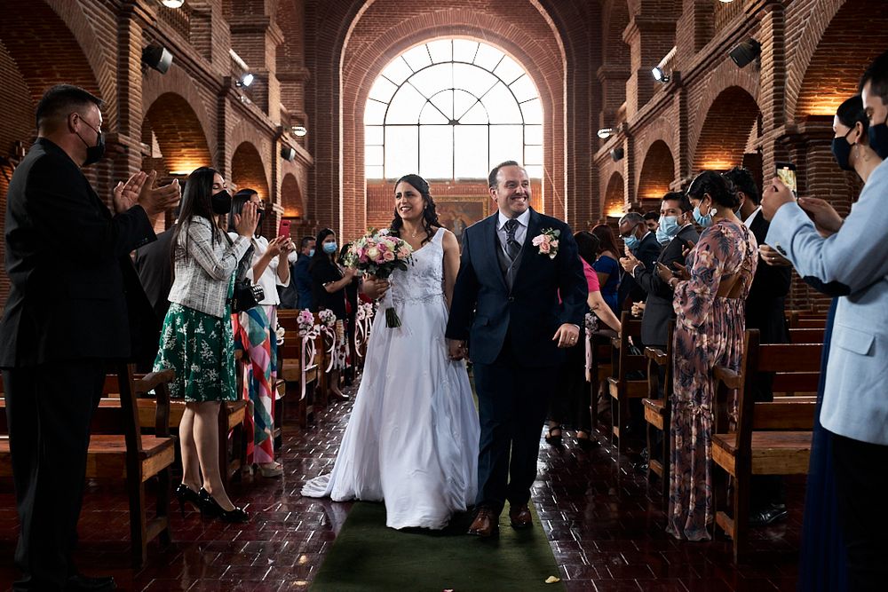 A couple walks down a grand church aisle with arched windows while guests look on.