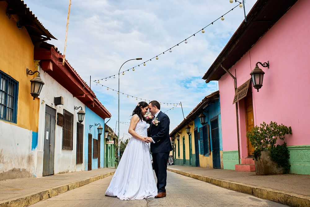 Newlyweds pose in colorful street with pastel buildings during wedding photoshoot.