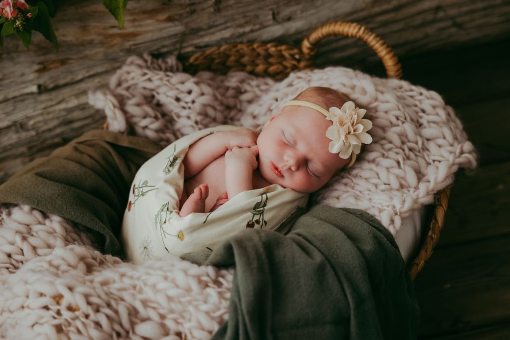 Peaceful sleeping newborn with white flower headband resting in woven basket.