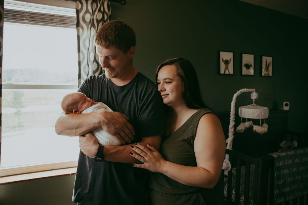 Parents stand by window while holding and admiring their sleeping newborn baby.