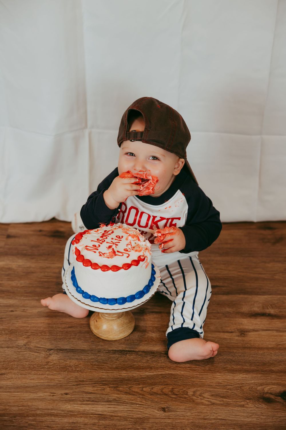 Baby in baseball outfit enjoying first birthday cake smash with blue and red frosting.