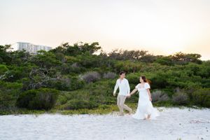 Couple walking on white sand beach with lush greenery and buildings in background.