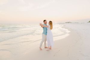 Couple in white and blue outfits dancing on a beach at sunset.