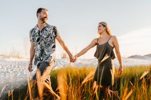 Couple holding hands while walking through golden grass field at sunset with mountains in background.