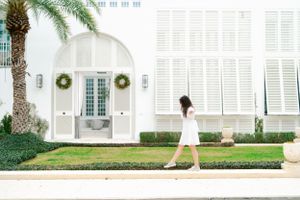 Woman in white dress walking past elegant white Mediterranean-style building with arched doorway and palm trees.