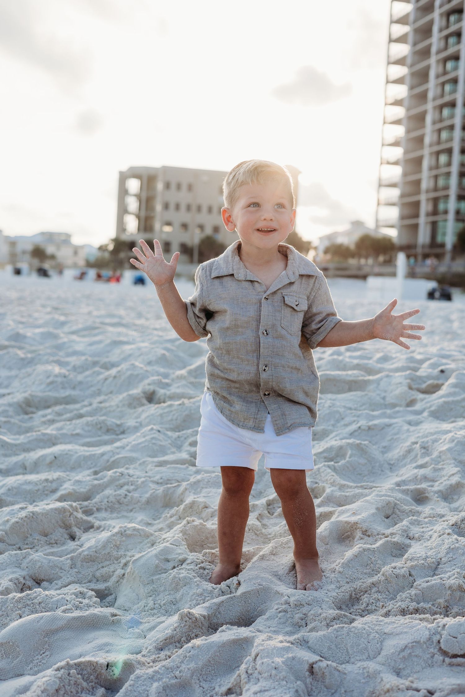 Happy toddler exploring and playing in beach sand with buildings in background.