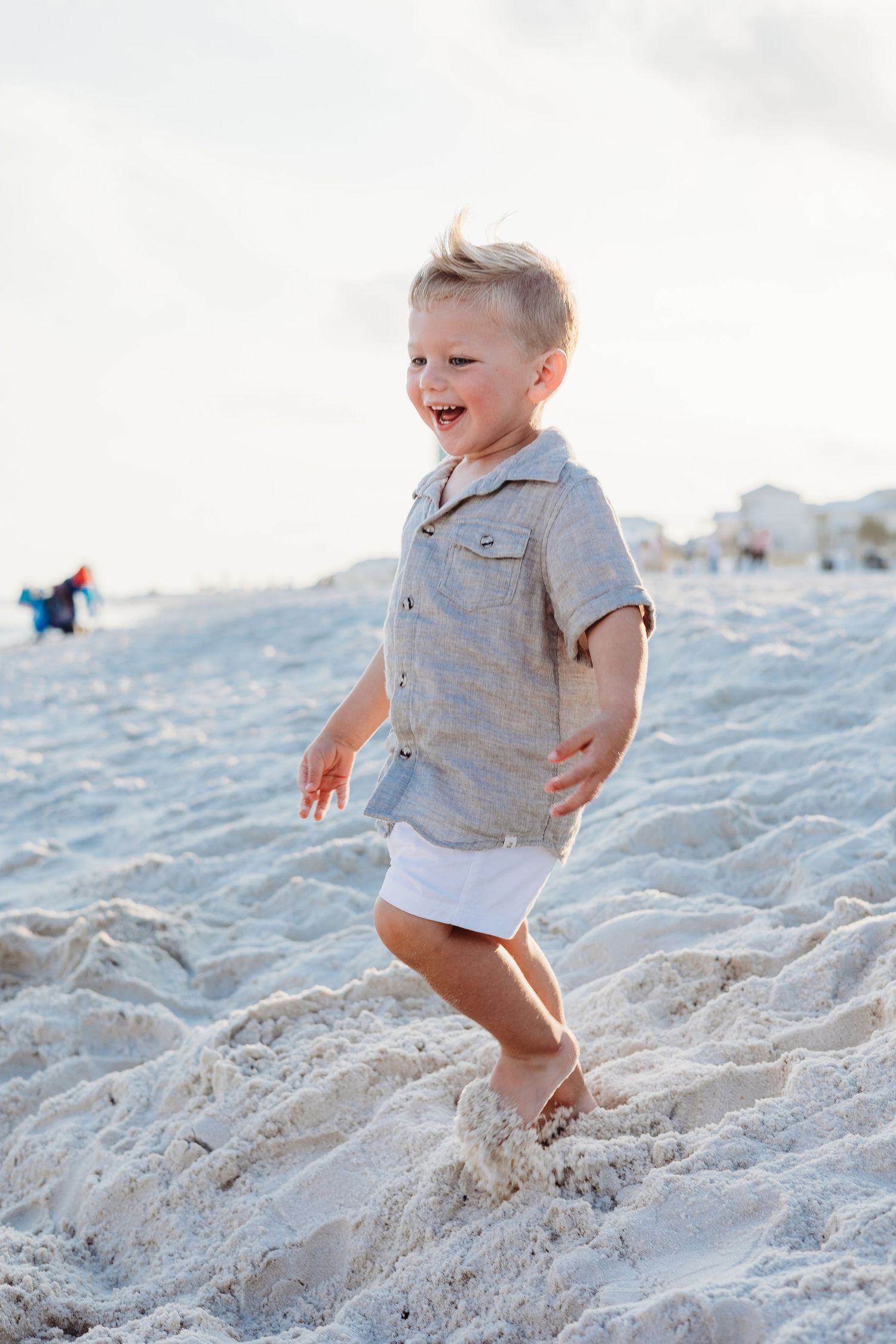 Toddler in grey shirt playing and jumping in white beach sand.
