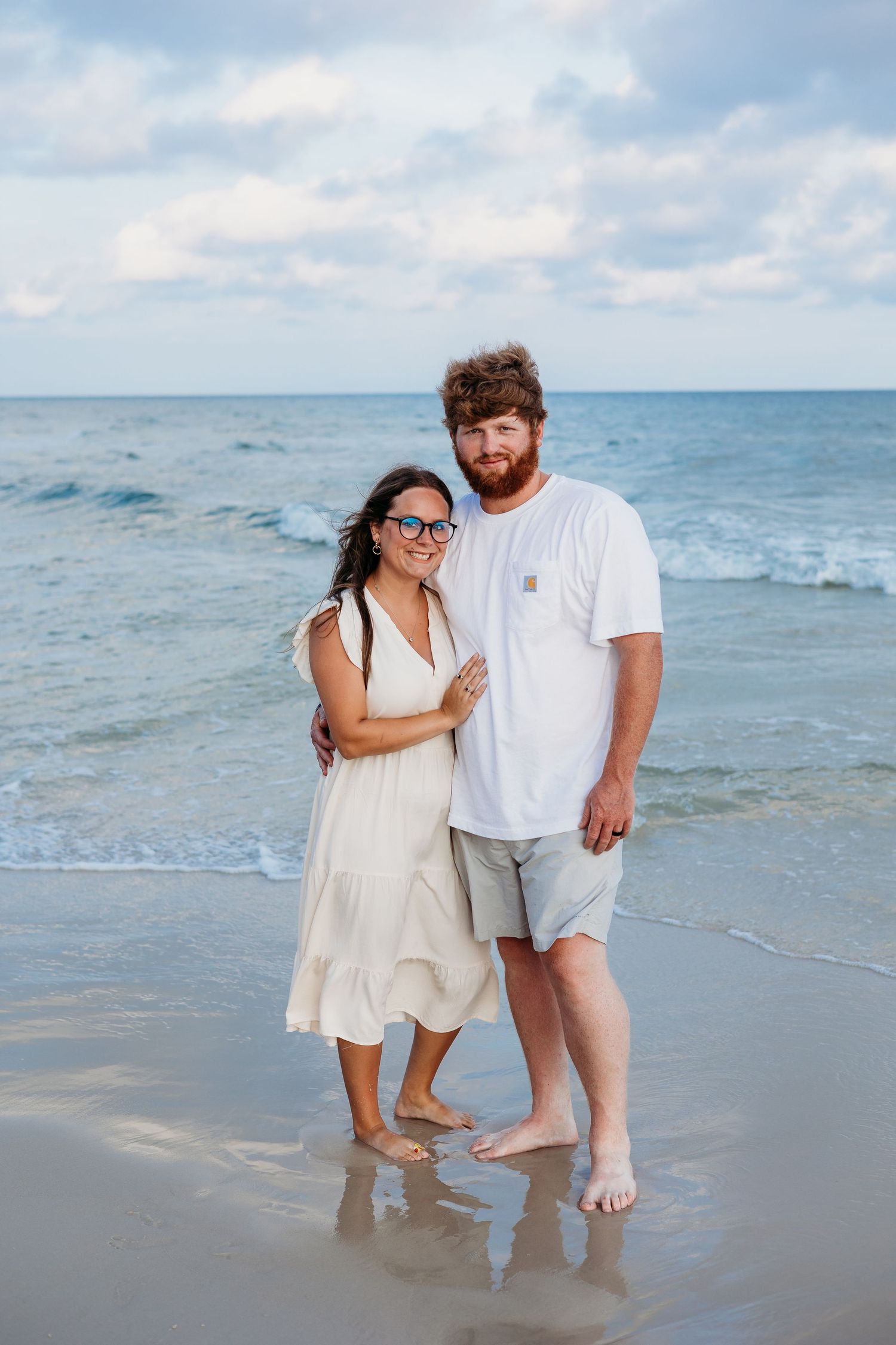 A couple dressed in white clothing poses playfully together at the water's edge on a beach.