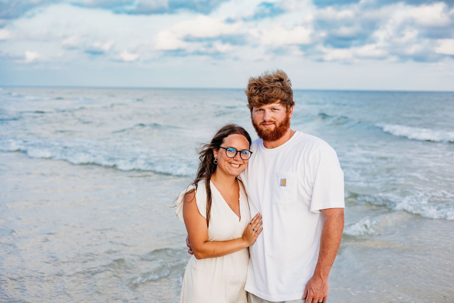 A couple in white outfits shares romantic moments while walking along a sunny beach with waves in the background.