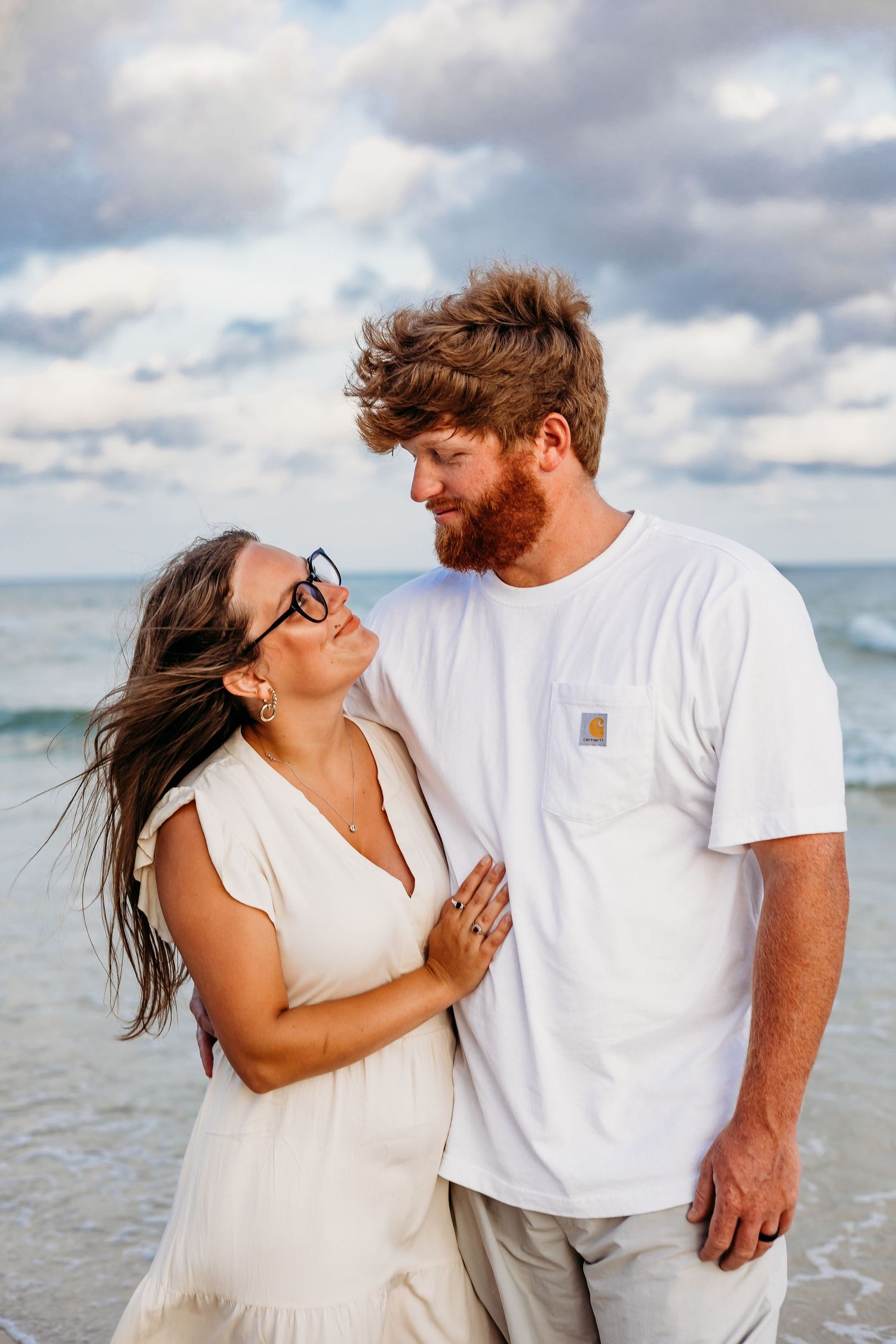 A couple in coordinated white outfits embraces and laughs together on a beach with cloudy skies.