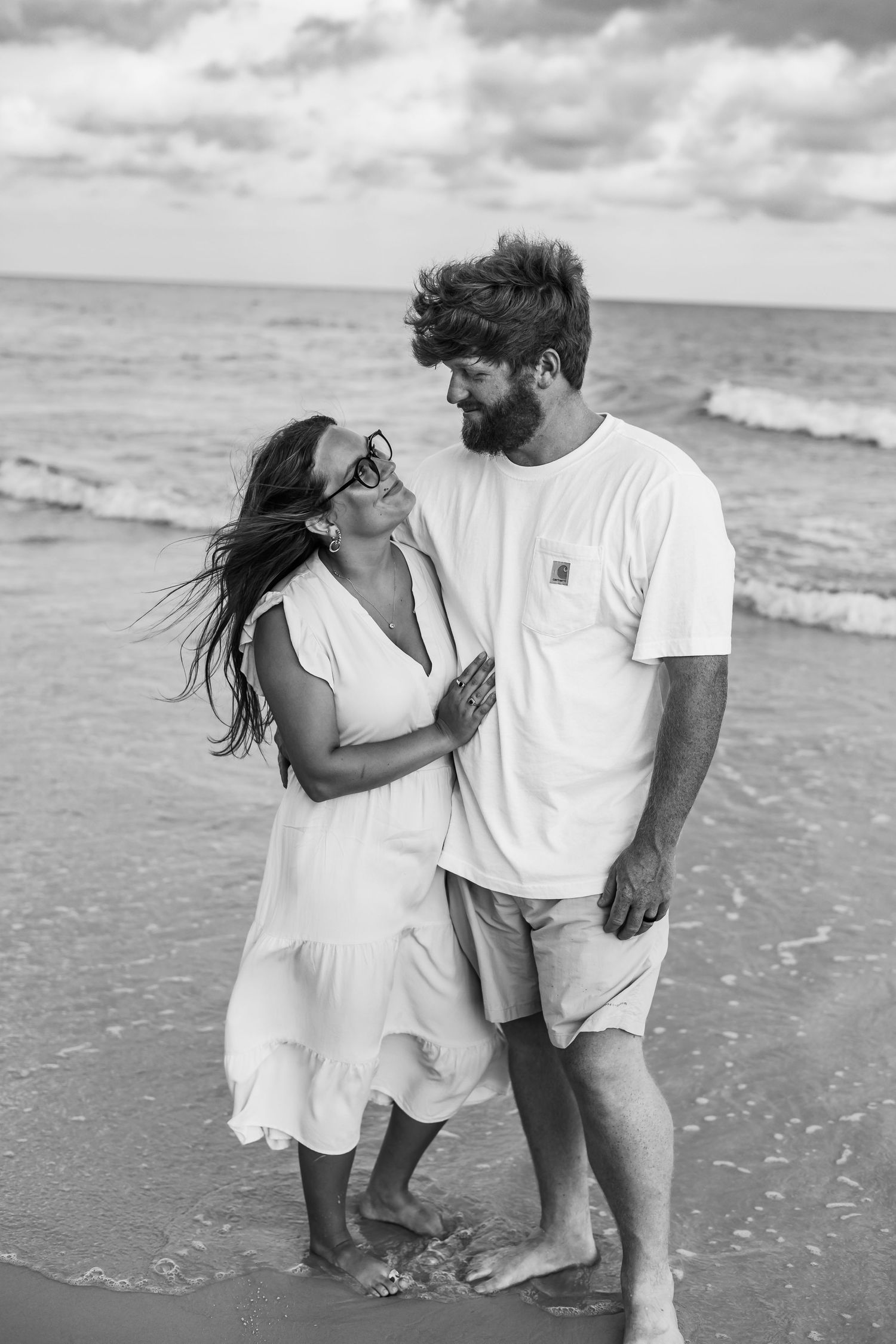 Black and white photo of a couple in white outfits sharing intimate moments on a beach.