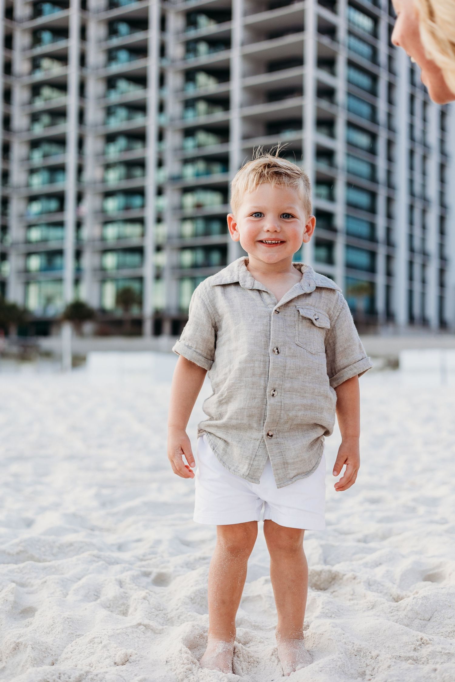 A small child in a gray button-up shirt plays in white sand with high-rise buildings in the background.