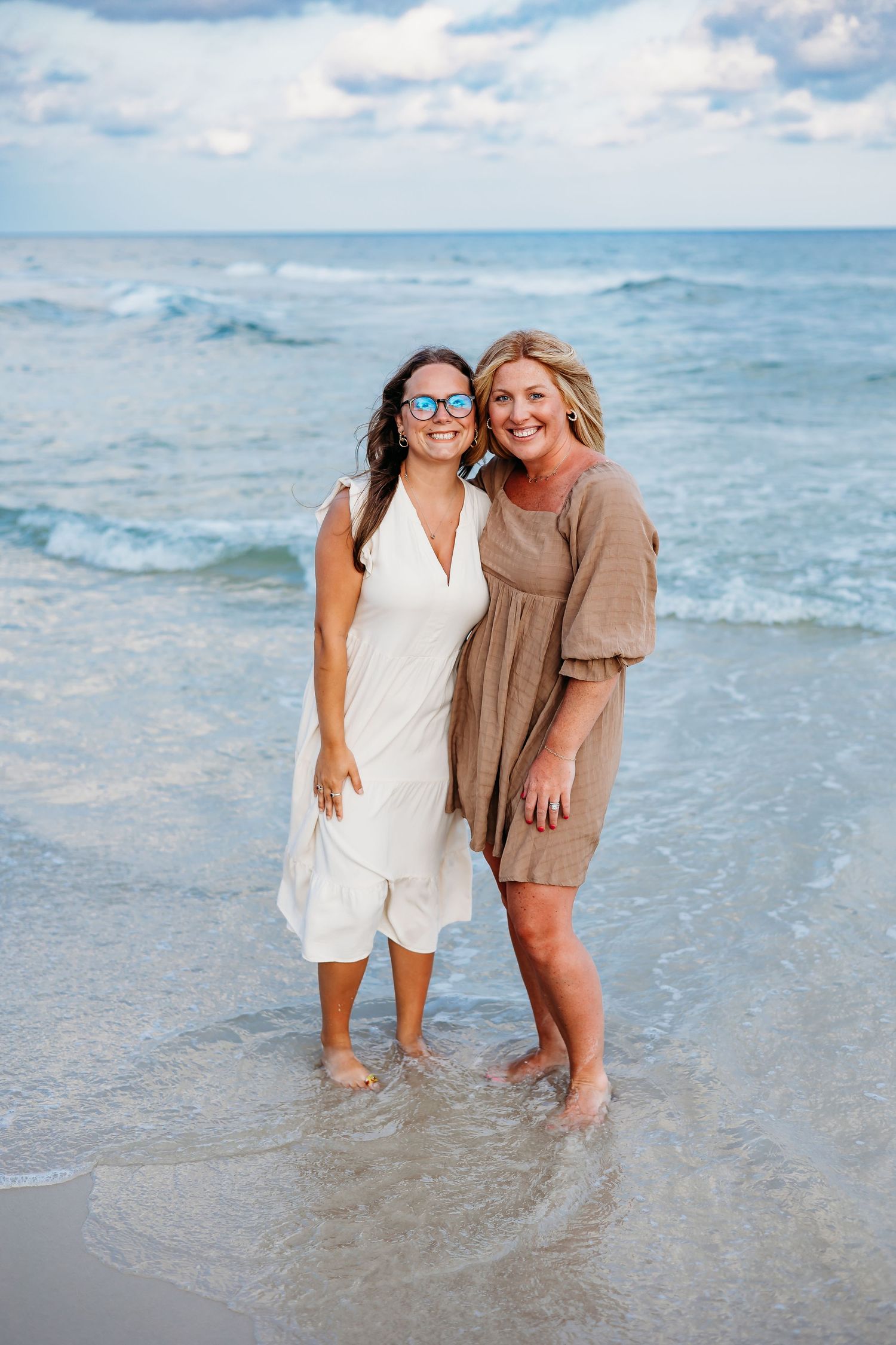 Two friends enjoying a sunny day at the beach wearing summer dresses with waves crashing behind them.