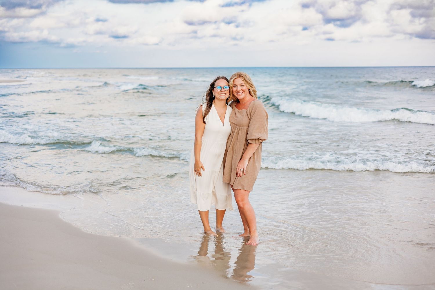 Two people standing together on a sandy beach with gentle waves and blue sky in the background.