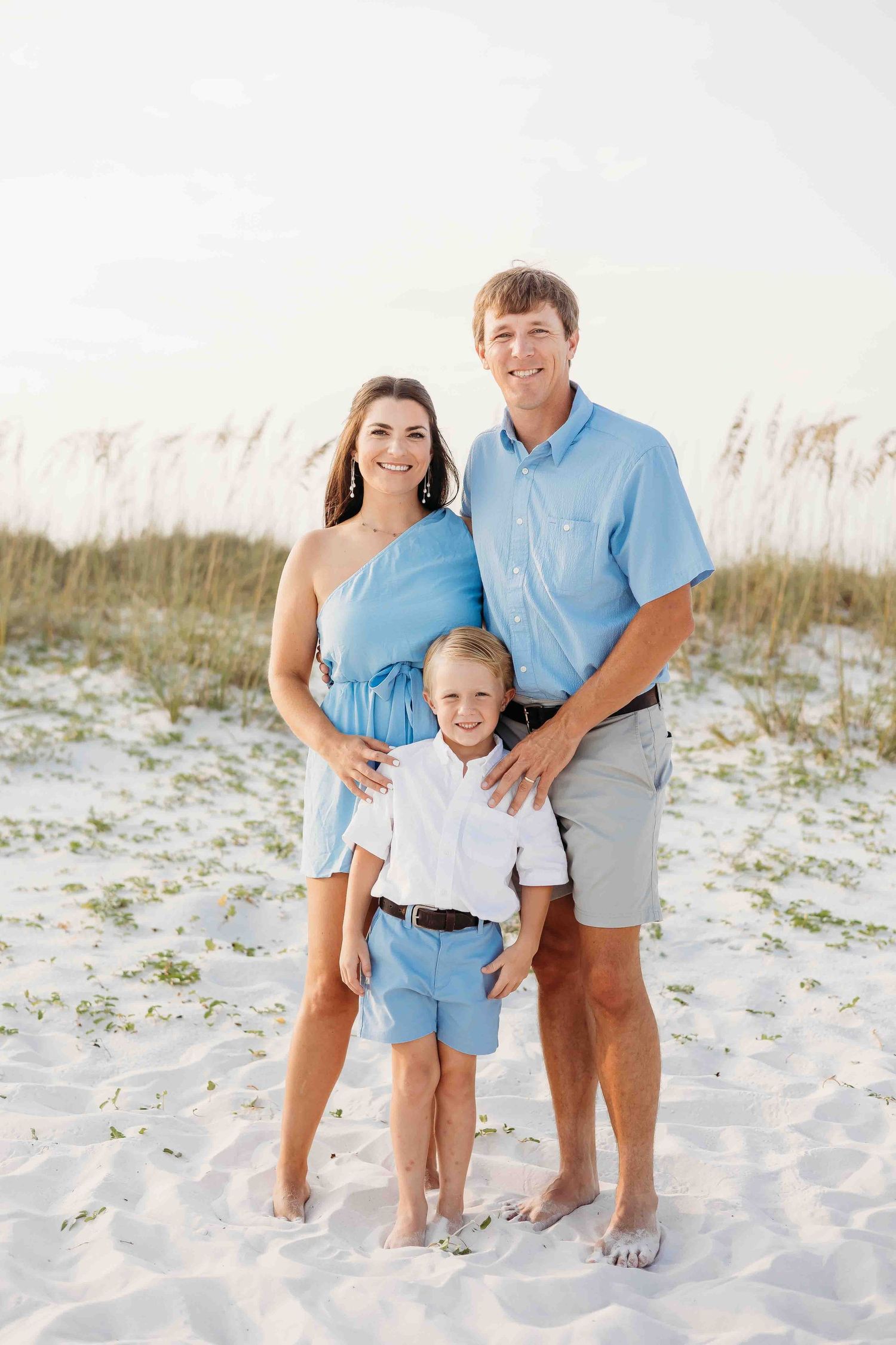 A family wearing coordinated blue and white outfits poses together on a sandy white beach at sunset.