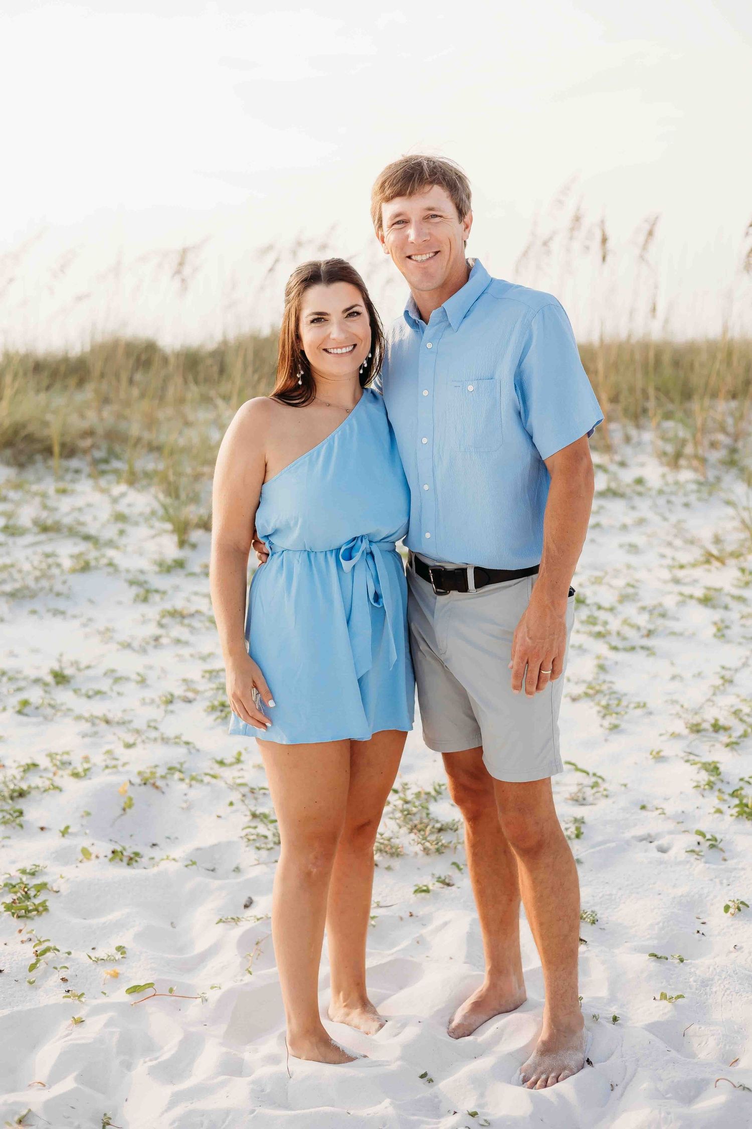 A couple in matching light blue outfits stands barefoot on pristine white sand dunes.