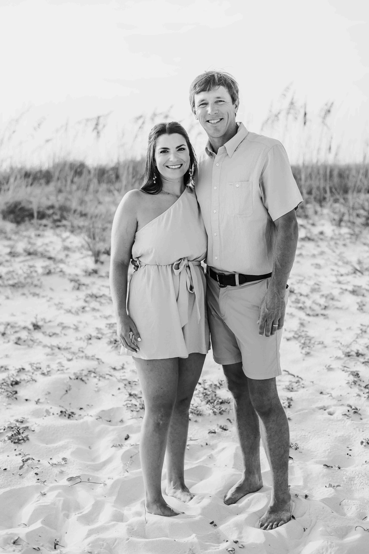 Black and white photo of a couple posing together on beach sand with sea oats in background.