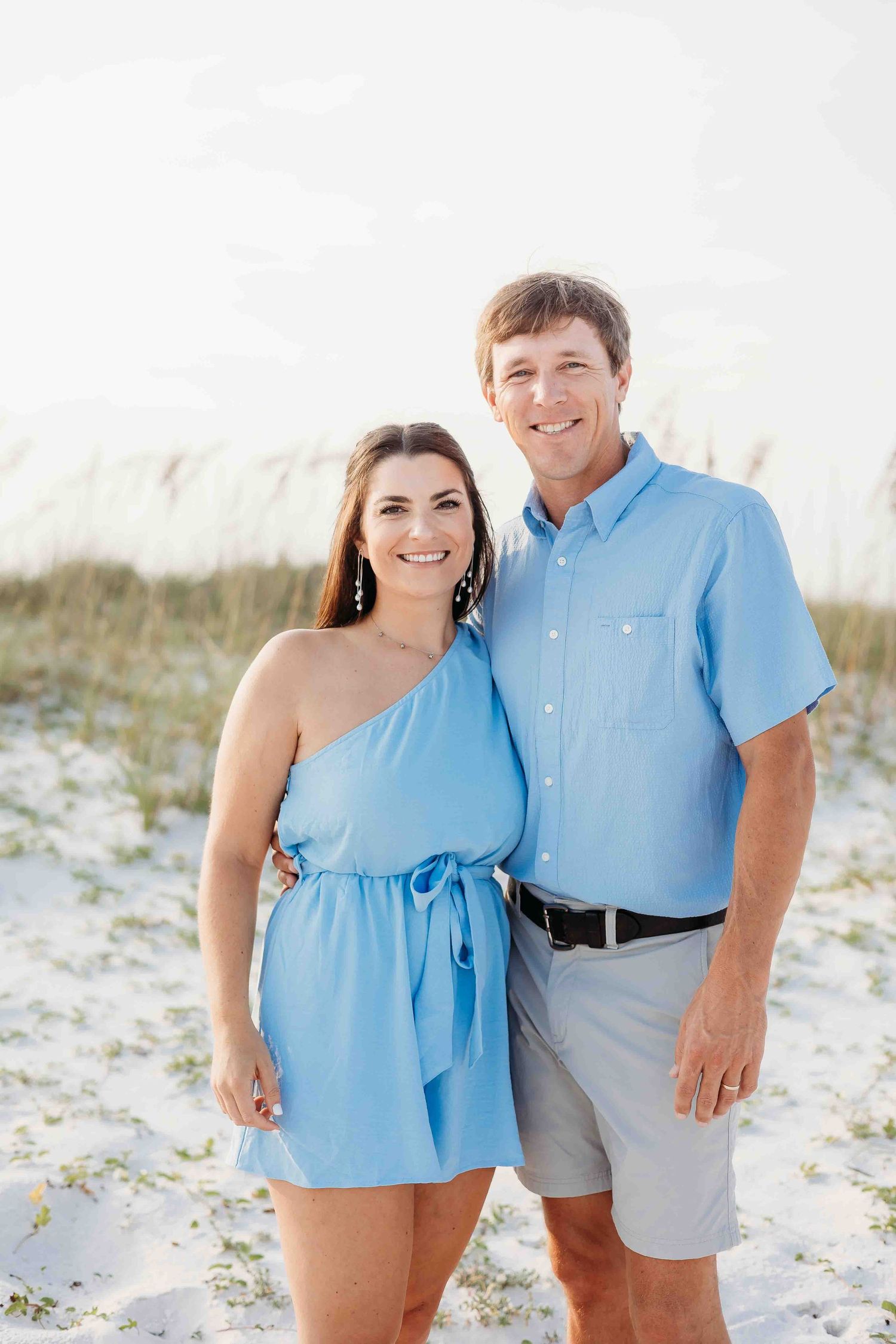 A couple in coordinated blue outfits smiles while standing together on a sandy beach.
