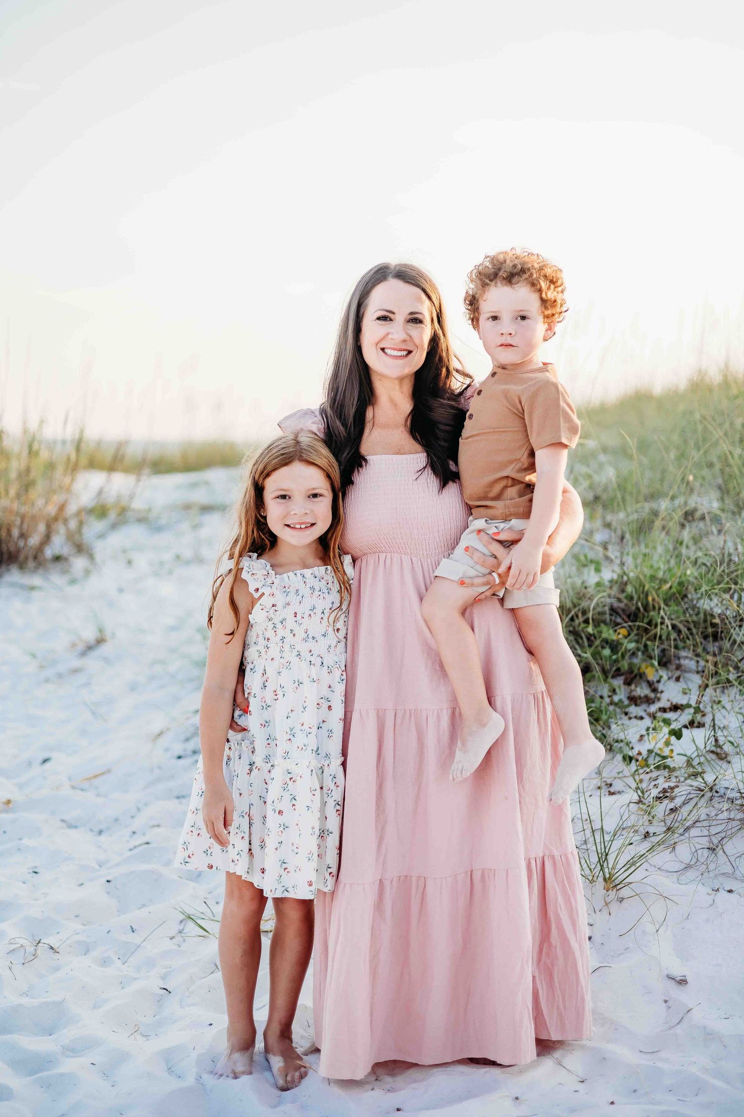 Three people in pink and floral dresses pose together on a beach with sand dunes in the background.