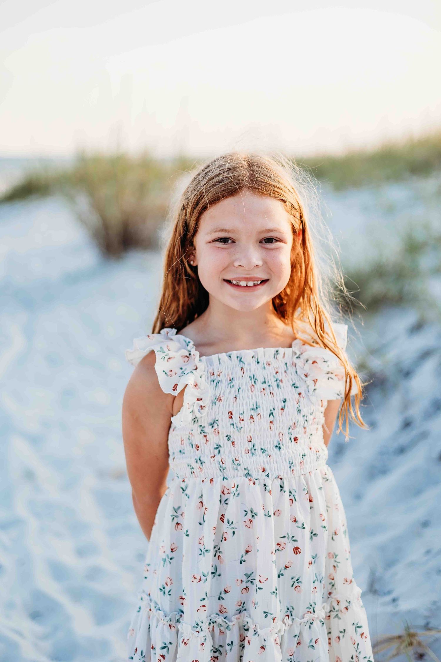 Young girl in a white floral dress smiles during a beach photo session.