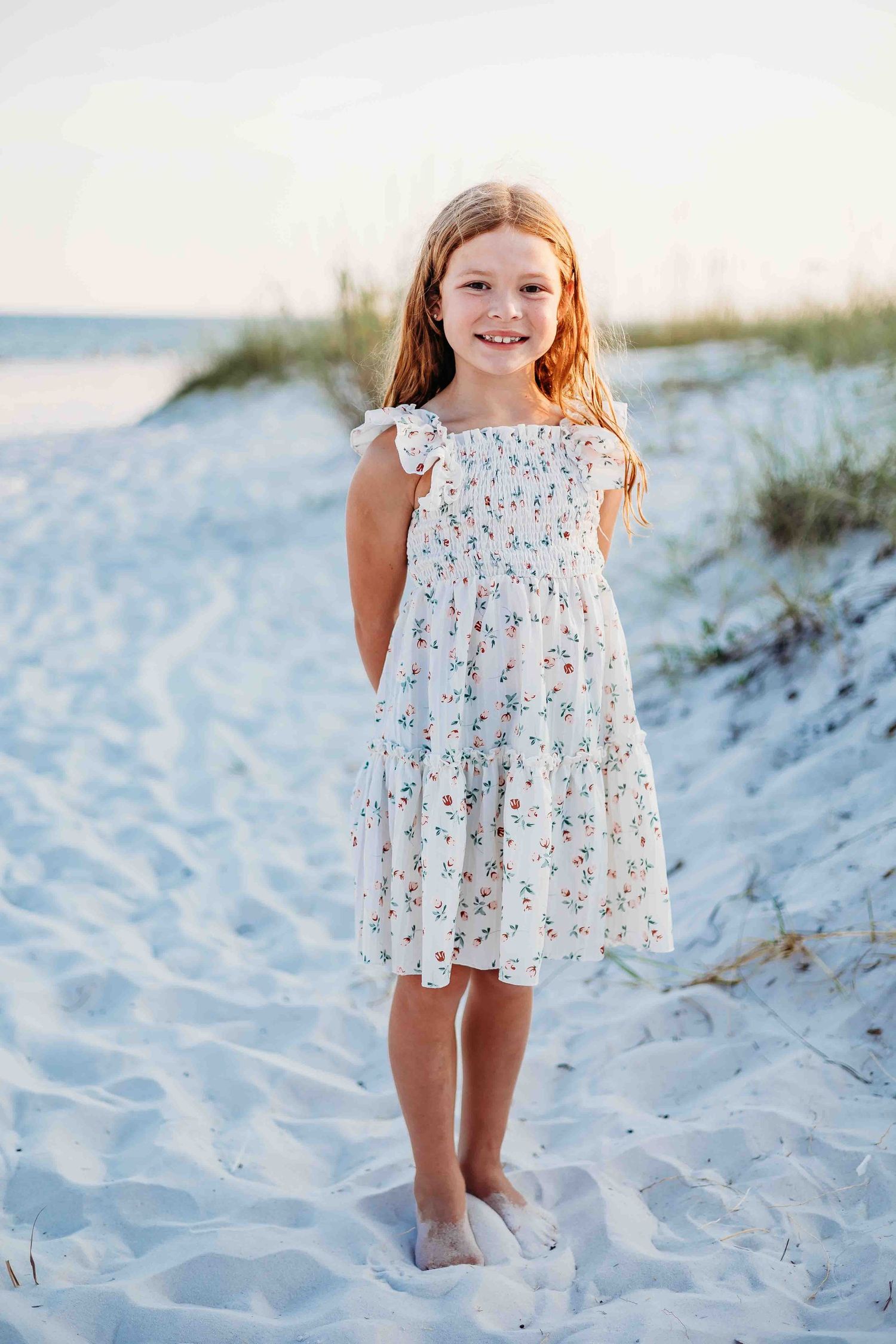 Child wearing a white floral dress poses playfully on white beach sand.