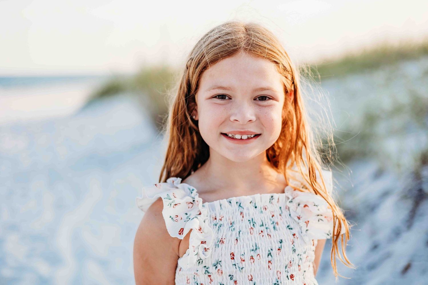 A floral sundress worn at a sunny beach with white sand and coastal vegetation.