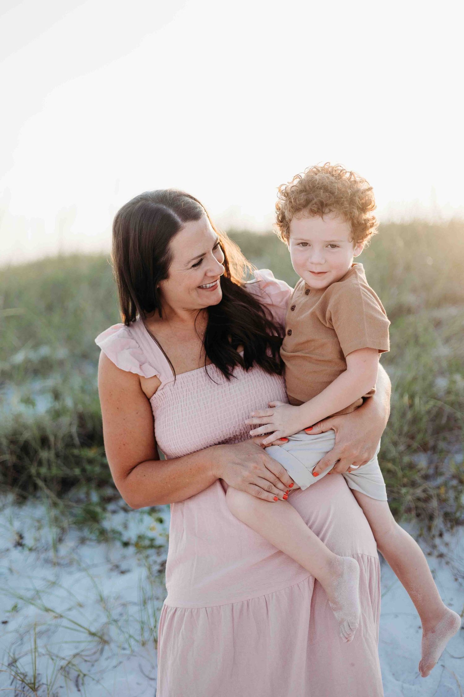 Tender family moment captured in soft pink tones against sandy beach backdrop.