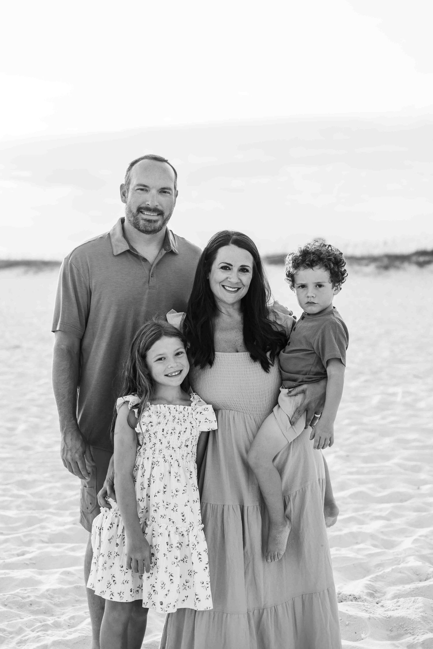 Family wearing coordinated outfits poses together on a sunny beach in black and white.