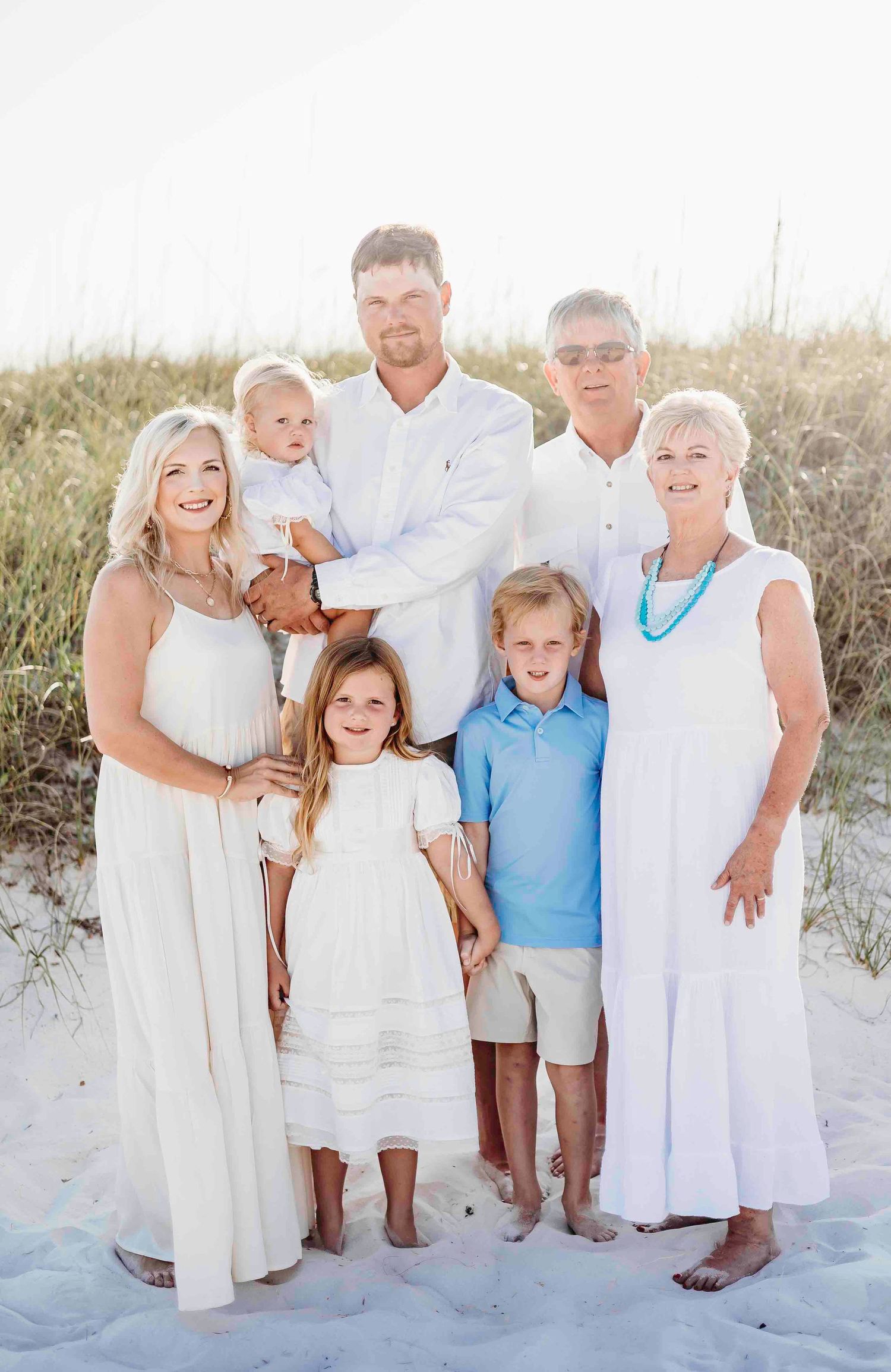 Extended family wearing white clothing poses together on white sand beach at sunset.