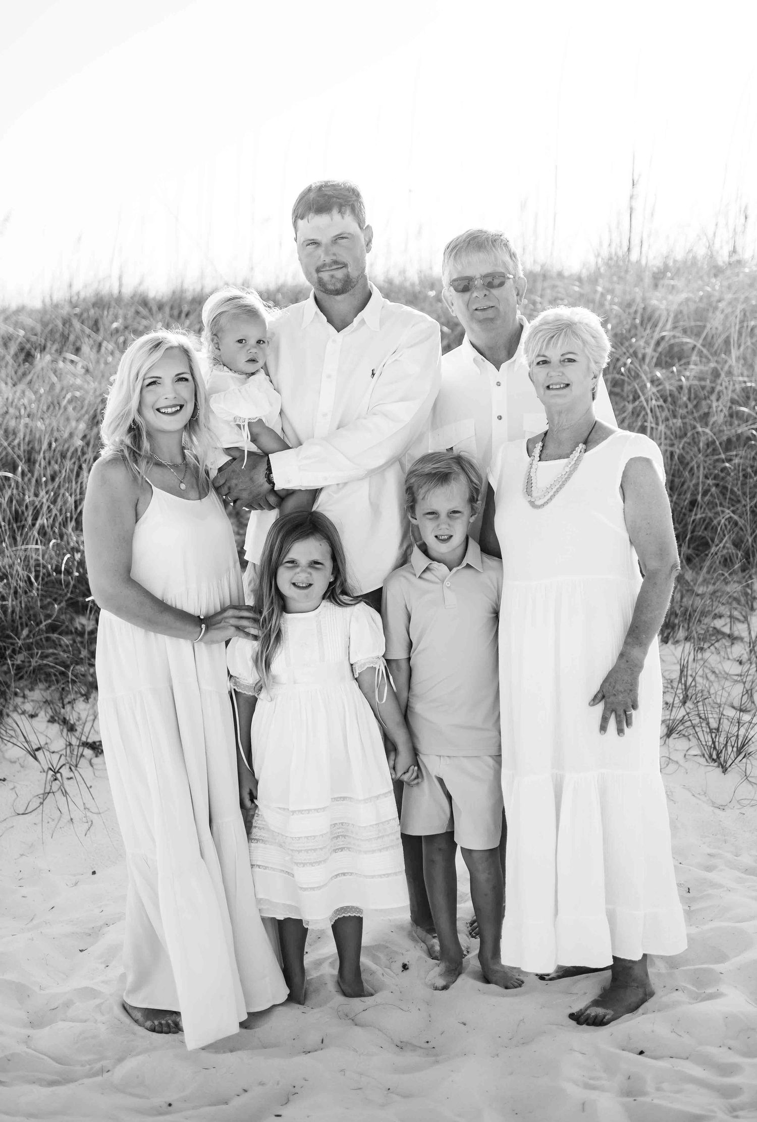 Multi-generational family group photo on beach wearing coordinated white outfits.