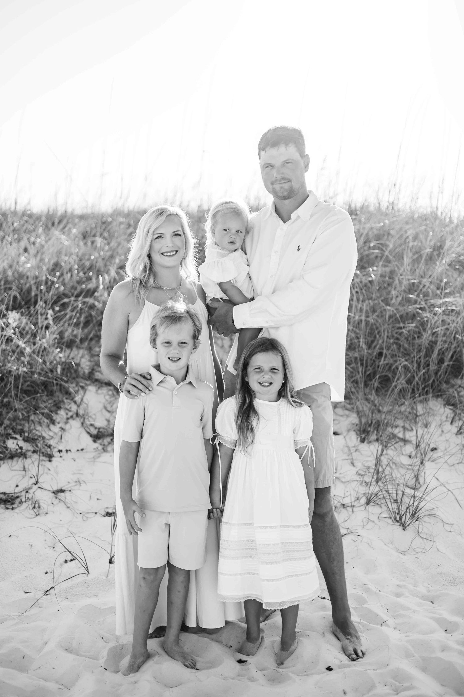 Family group poses casually on sandy beach dunes in light summer clothing.