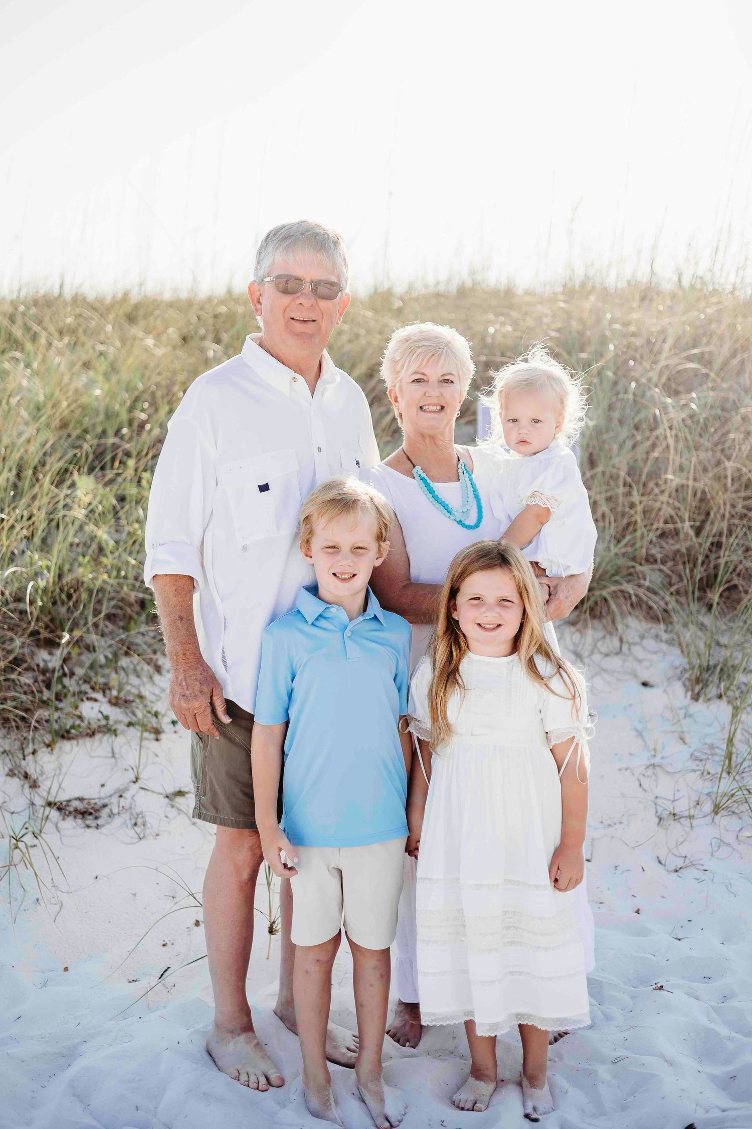 Extended family poses together on beach sand wearing coordinated white and blue outfits.