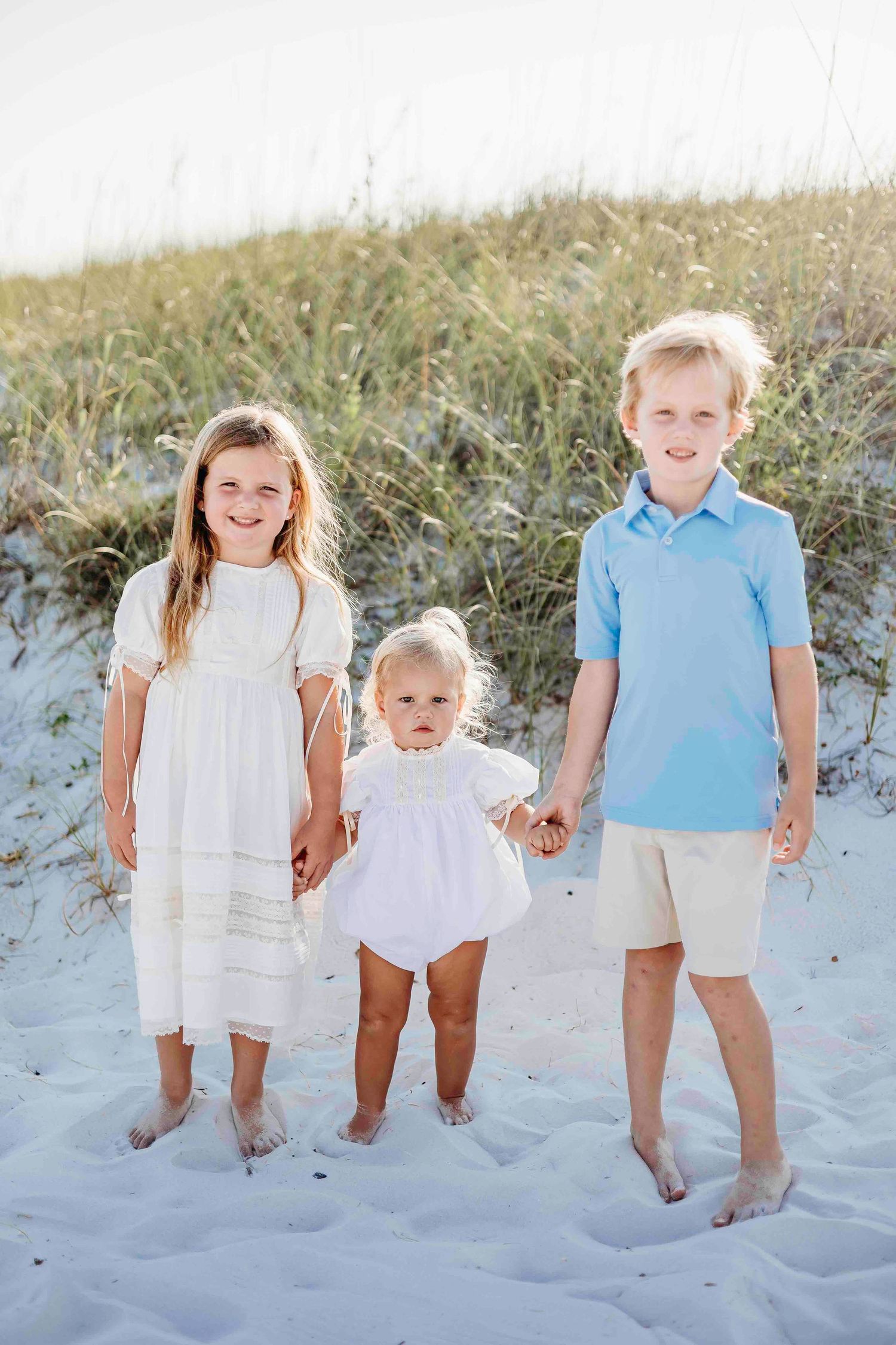 Children in coordinating white and blue outfits standing together on white sand beach.