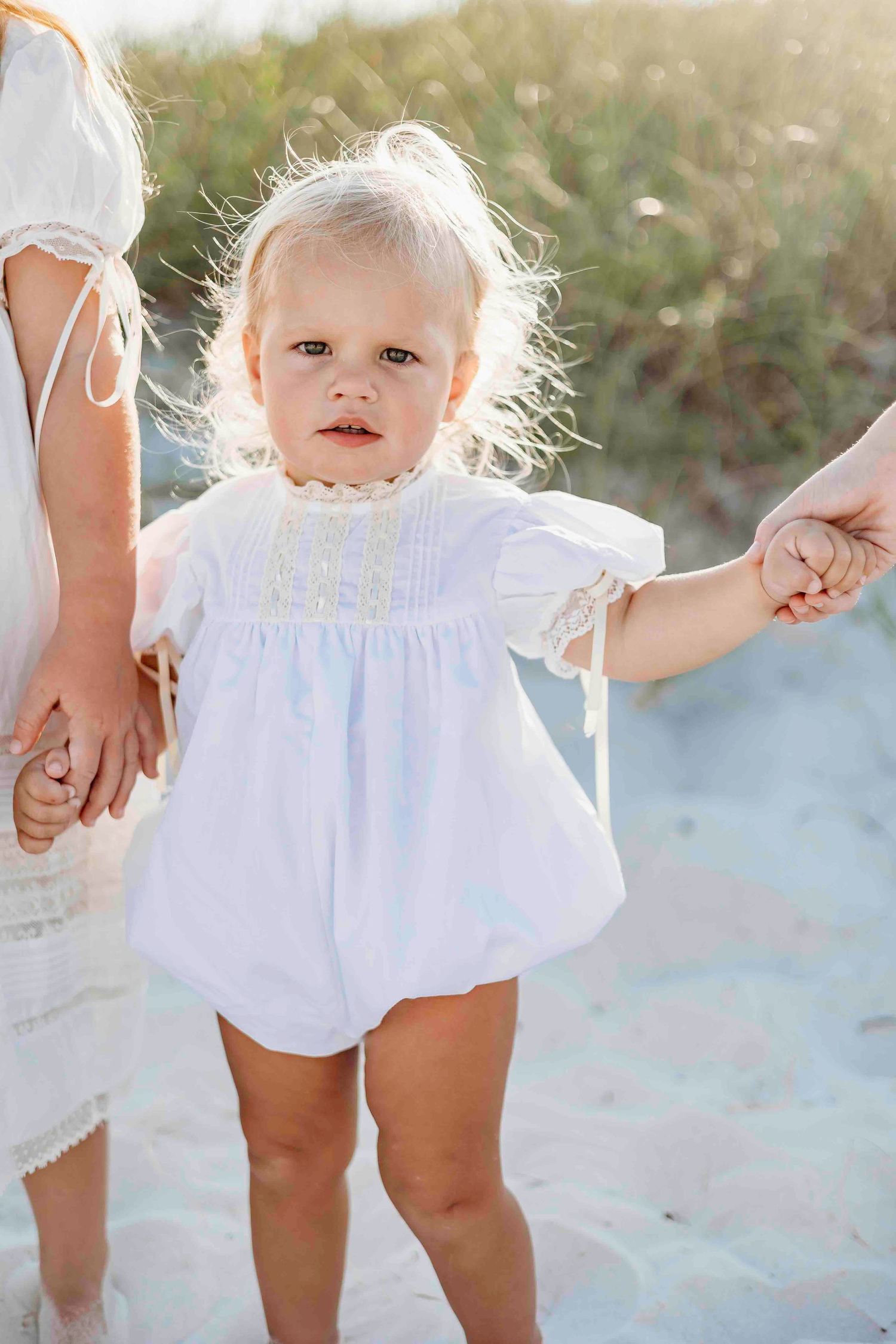 Close-up view of children in white summer dresses standing on beach sand.