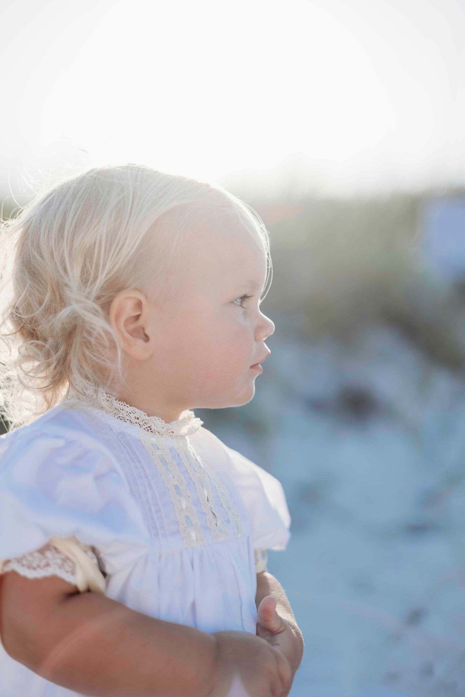 Close-up profile view of a toddler wearing a white lace dress at the beach.