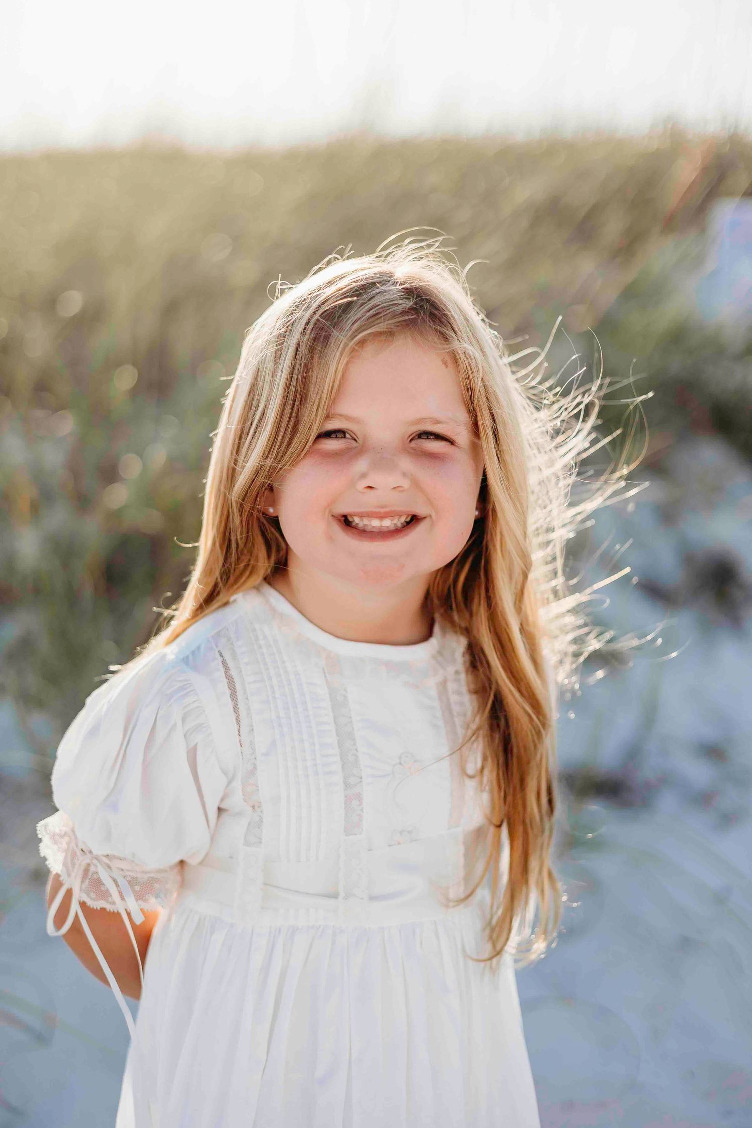 A child in a white vintage dress smiles while playing in sand dunes.