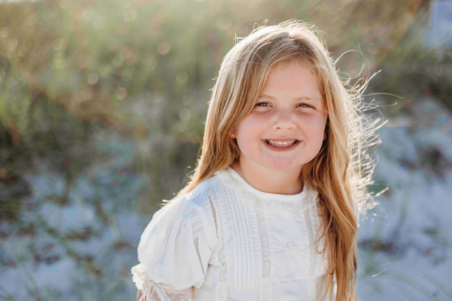 A happy child wearing a white dress poses on sandy dunes at the beach.