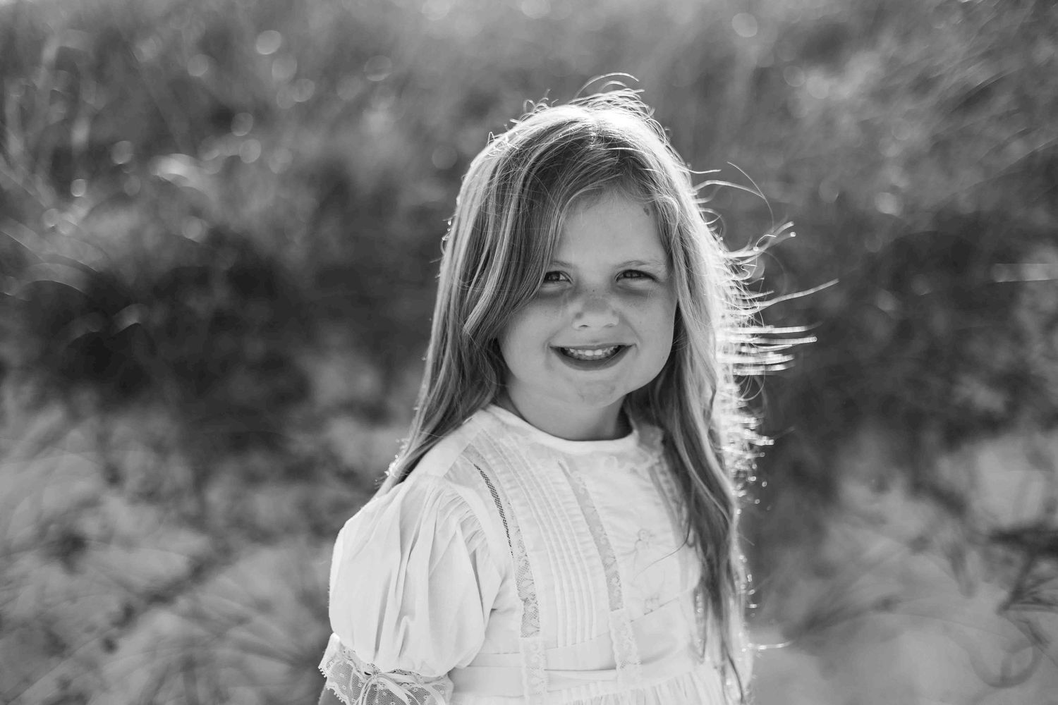 Black and white portrait of a child smiling in a white dress outdoors.