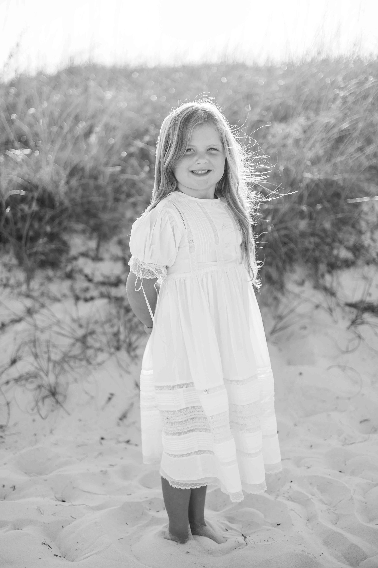 Black and white photo of a child in vintage dress standing on beach sand.