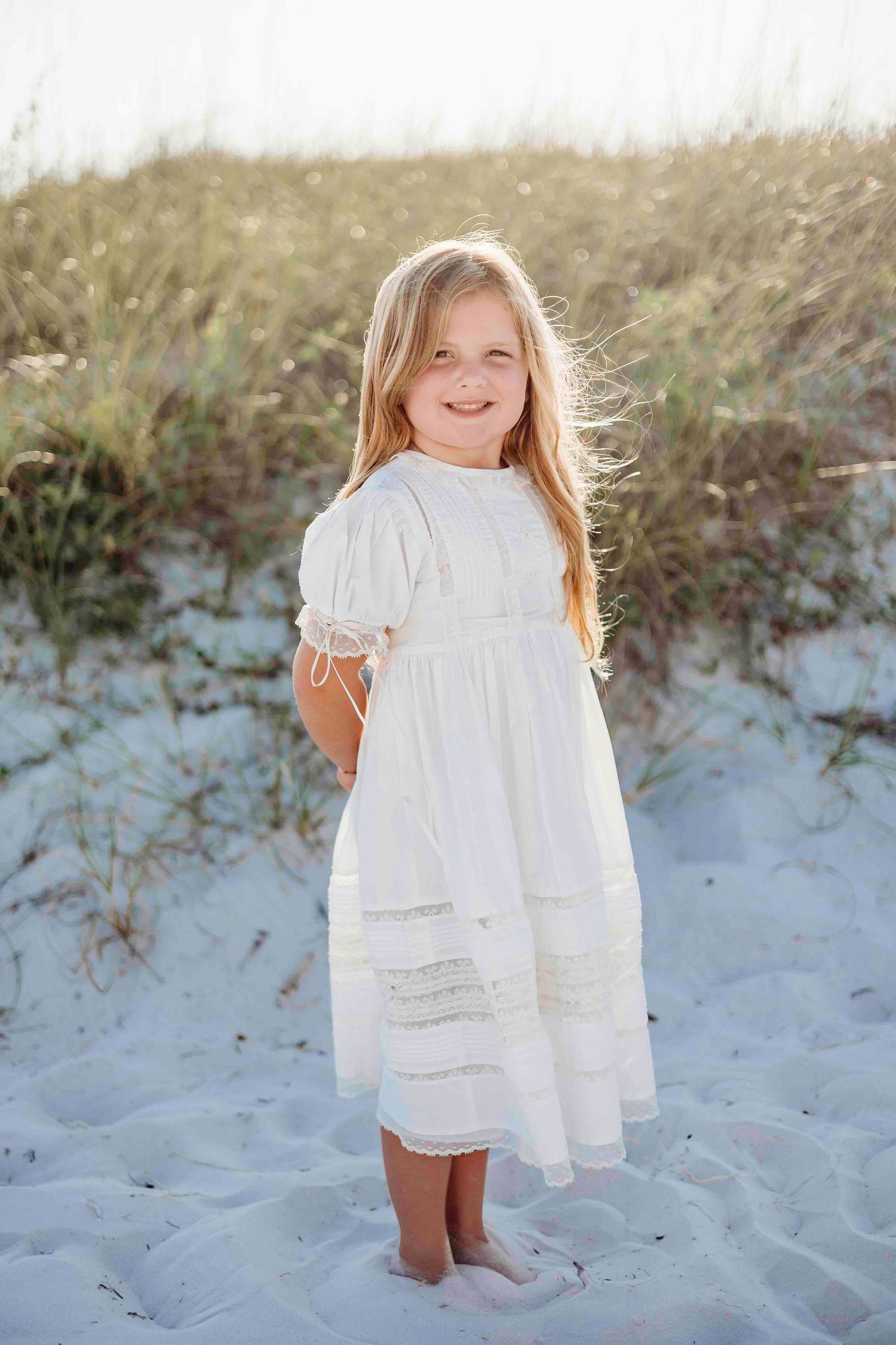 A child in white dress stands barefoot on sandy beach dunes.